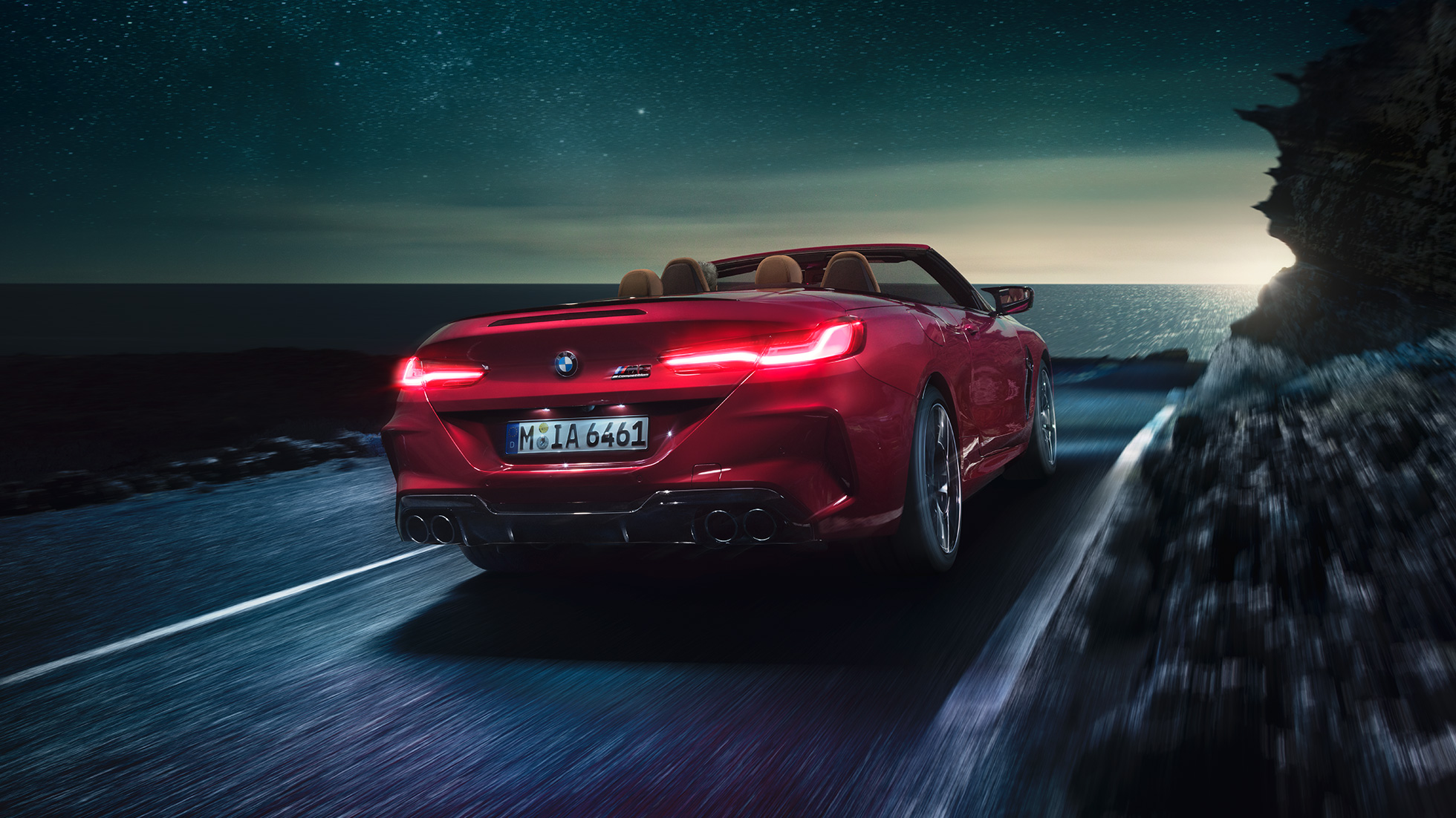BMW M8 Competition Кабриолет, вид сзади с поворотом в три четверти. BMW M8 Competition Кабриолет в Motegi Red металлик с открытым мягким верхом, вид сзади с поворотом в три четверти, езда ночью по прибрежной дороге.