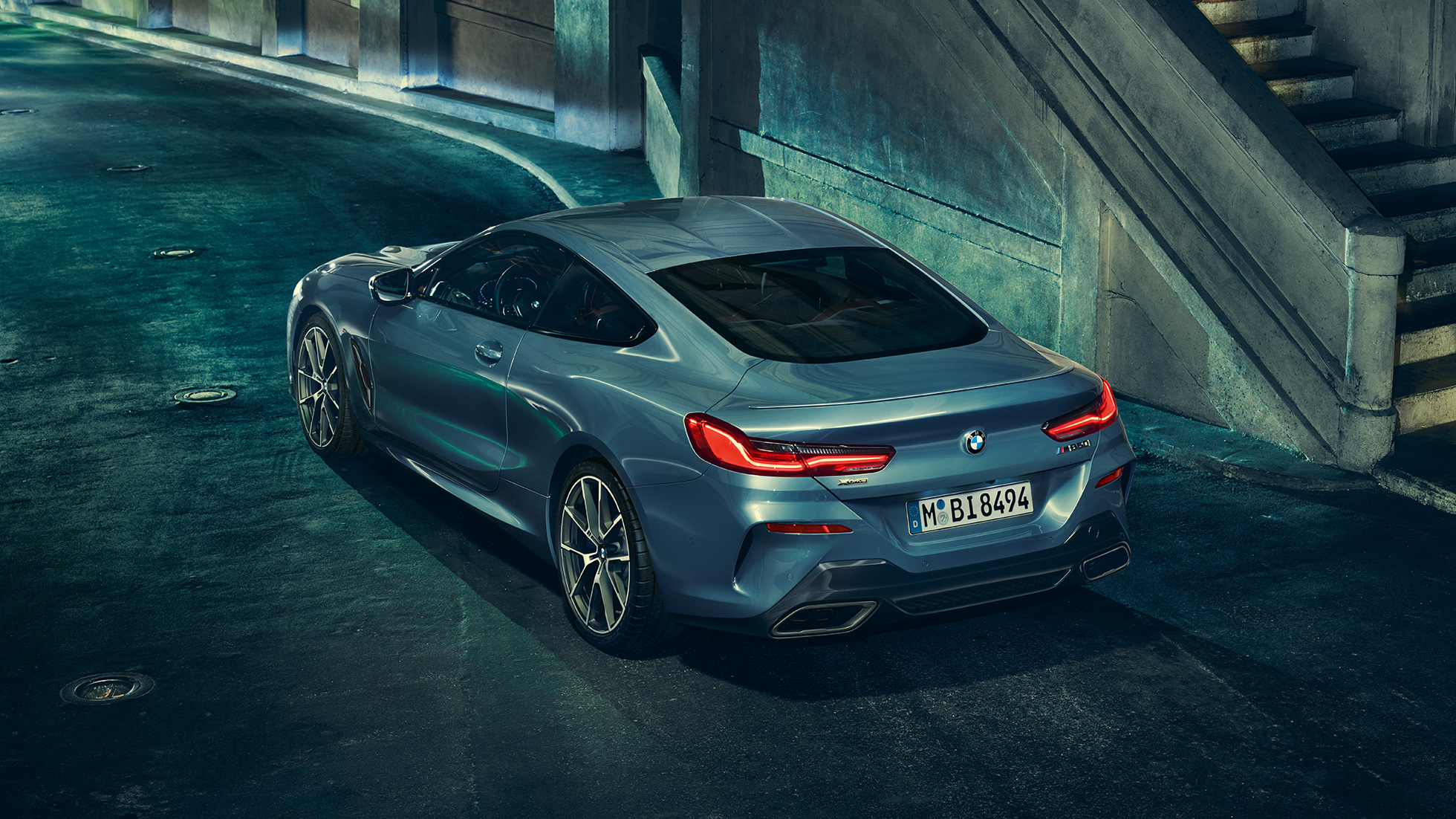 BMW M850i xDrive Coupé, вид сзади с поворотом в три четверти слева. BMW M850i xDrive Coupé в Barcelona Blue металлик, вид сзади с поворотом в три четверти слева, стоит ночью.